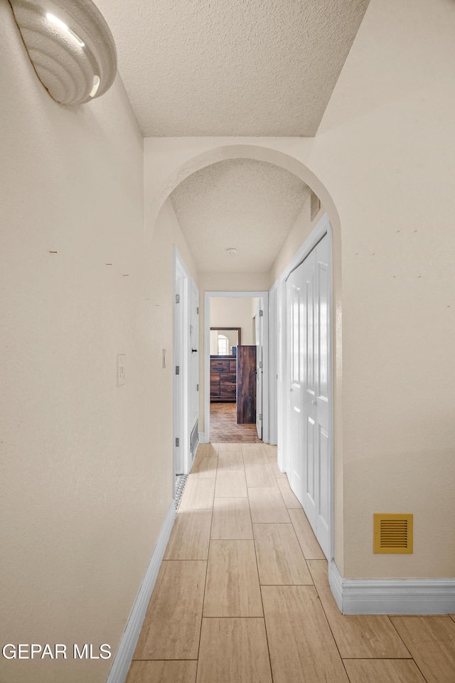 hall with a textured ceiling, arched walkways, baseboards, and light wood-type flooring