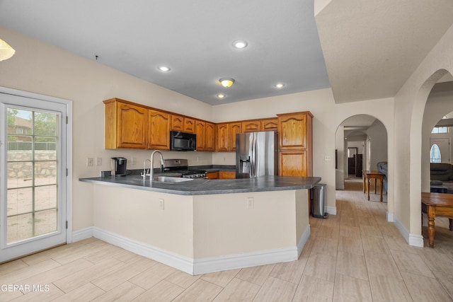 kitchen with stainless steel appliances, arched walkways, a peninsula, brown cabinetry, and dark countertops