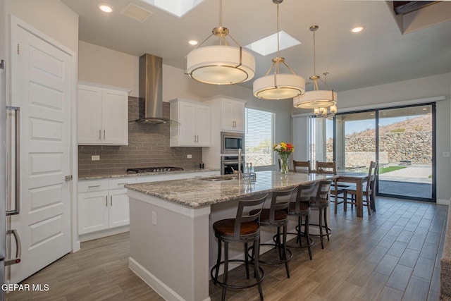 7389 Cimarron Gap Dr, El Paso, TX 79911 - kitchen with wall chimney range hood, appliances with stainless steel finishes, backsplash, a center island with sink, and wood finished floors