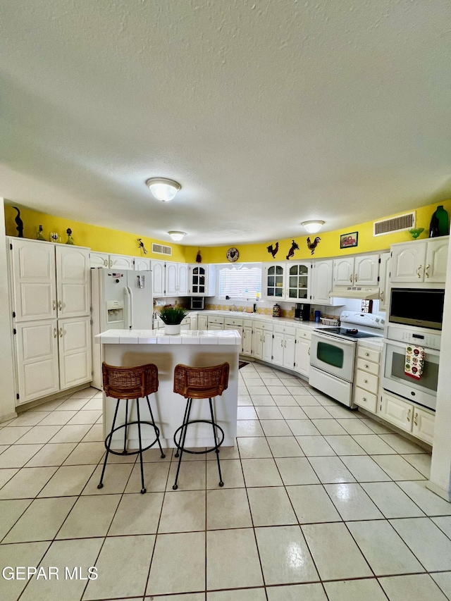 16899 Old Butterfield Trl, El Paso, TX 79938 - kitchen featuring white appliances, tile countertops, a breakfast bar, white cabinetry, and under cabinet range hood