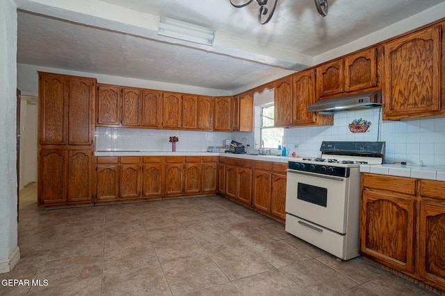 363 Ontiveros St, El Paso, TX 79932 - kitchen featuring white gas range oven, under cabinet range hood, brown cabinets, decorative backsplash, and tile countertops