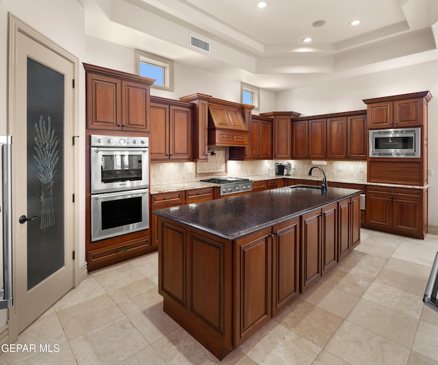 6572 Tuscany Ridge Dr, El Paso, TX 79912 - kitchen with appliances with stainless steel finishes, a tray ceiling, premium range hood, tasteful backsplash, and a kitchen island with sink