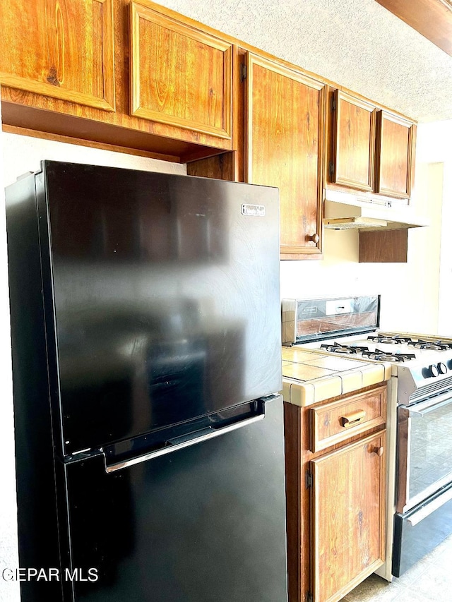 2520 San Jose Ave Unit 2D, El Paso, TX 79930 - kitchen with freestanding refrigerator, gas range gas stove, tile countertops, under cabinet range hood, and brown cabinets
