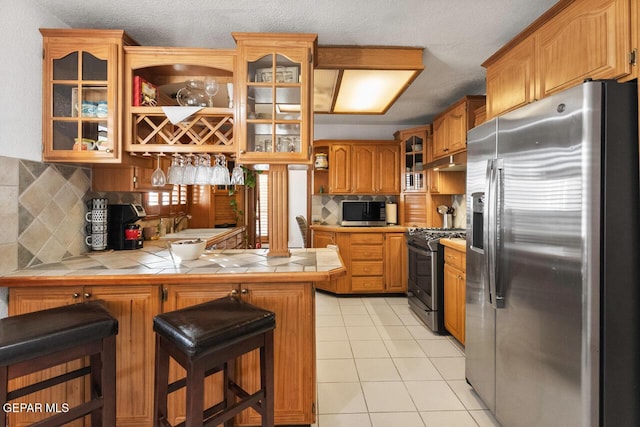 612 Firedale Ln, El Paso, TX 79912 - kitchen with appliances with stainless steel finishes, a peninsula, tile countertops, light tile patterned flooring, and under cabinet range hood