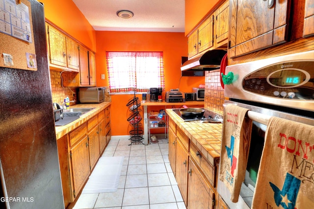 5108 Paris Ave, El Paso, TX 79924 - kitchen with under cabinet range hood, stainless steel appliances, backsplash, light tile patterned flooring, and brown cabinets