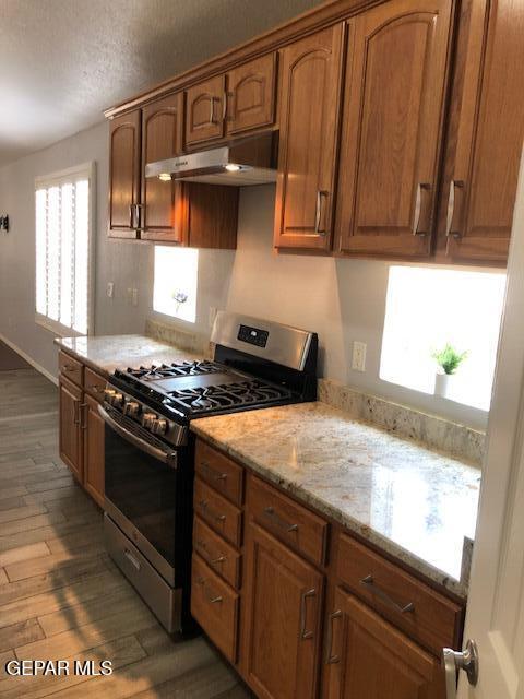 6392 Franklin Gate Dr, El Paso, TX 79912 - kitchen with stainless steel gas range, under cabinet range hood, light wood-style floors, light stone countertops, and brown cabinetry