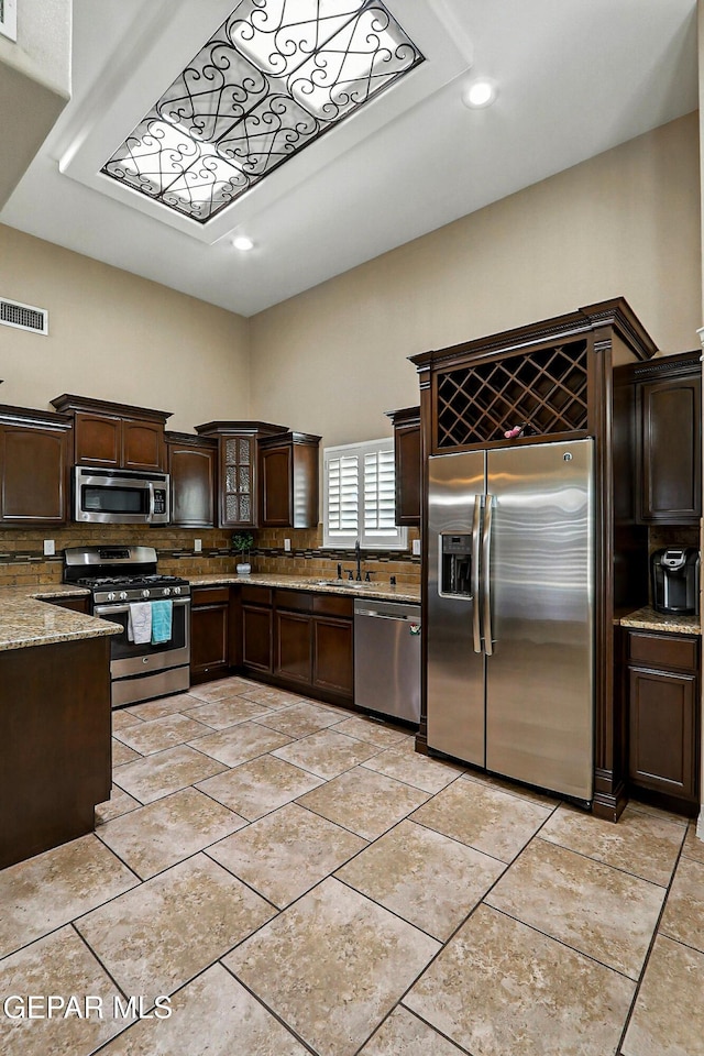 14552 Long Shadow Ave, El Paso, TX 79938 - kitchen with appliances with stainless steel finishes, decorative backsplash, dark brown cabinetry, light stone countertops, and glass insert cabinets