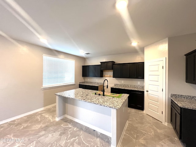3720 Hill Top Place, El Paso, TX 79938 - kitchen with light marble finish flooring, a center island with sink, light stone counters, and dark cabinetry