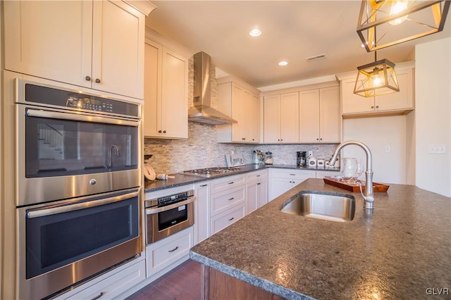 kitchen with decorative light fixtures, stainless steel appliances, sink, white cabinetry, and wall chimney range hood