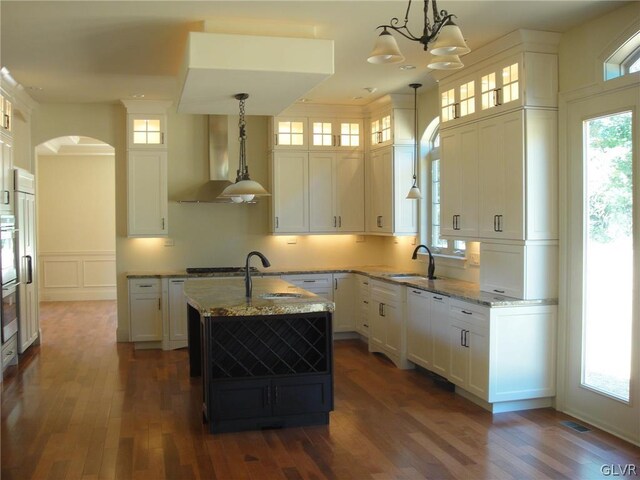 kitchen with sink, wall chimney range hood, a kitchen island with sink, light stone countertops, and decorative light fixtures