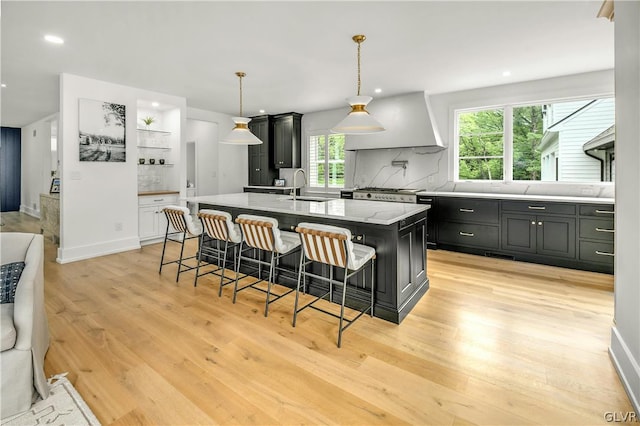 kitchen with an island with sink, sink, decorative backsplash, custom exhaust hood, and light hardwood / wood-style flooring