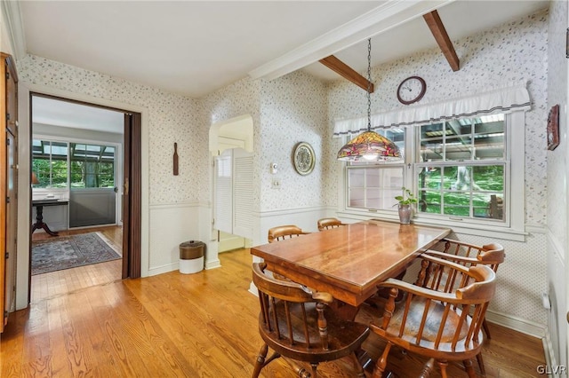 dining area featuring wallpapered walls, light wood finished floors, beam ceiling, and wainscoting