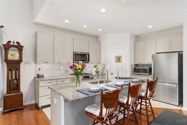 kitchen with light hardwood / wood-style flooring, appliances with stainless steel finishes, light stone counters, sink, and an island with sink