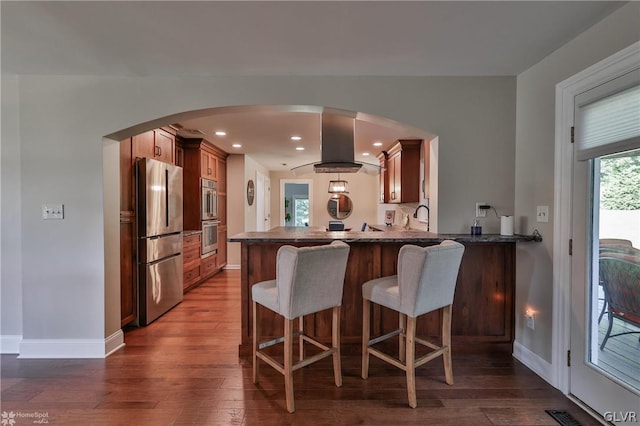 kitchen with appliances with stainless steel finishes, a breakfast bar area, kitchen peninsula, exhaust hood, and dark wood-type flooring