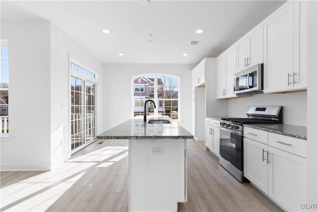 kitchen with stainless steel appliances, light hardwood / wood-style flooring, a center island with sink, and dark stone countertops