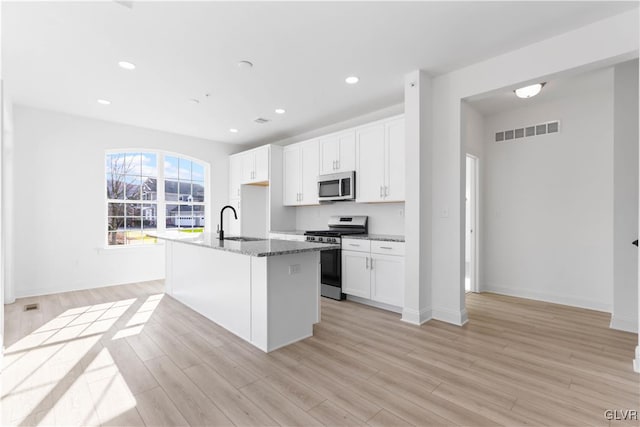 kitchen featuring white cabinetry, dark stone countertops, an island with sink, appliances with stainless steel finishes, and light wood-type flooring