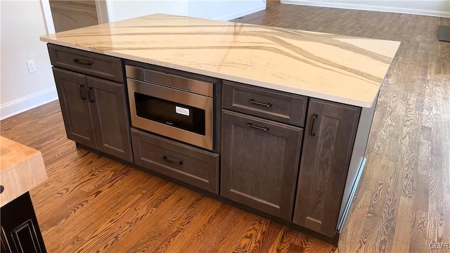 kitchen with dark brown cabinetry, stainless steel microwave, dark hardwood / wood-style floors, and light stone countertops