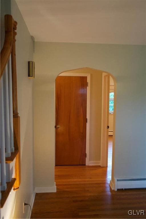 hallway with a baseboard heating unit, wood finished floors, arched walkways, and baseboards