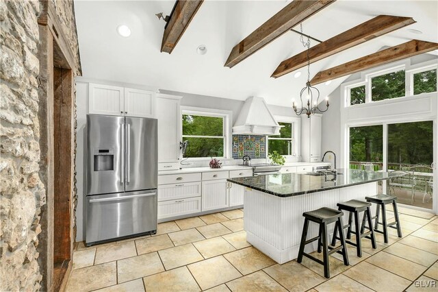 kitchen featuring an island with sink, premium range hood, stainless steel fridge with ice dispenser, pendant lighting, and white cabinets