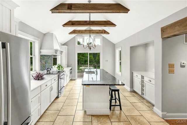 kitchen featuring wall chimney exhaust hood, beamed ceiling, an island with sink, and stainless steel appliances