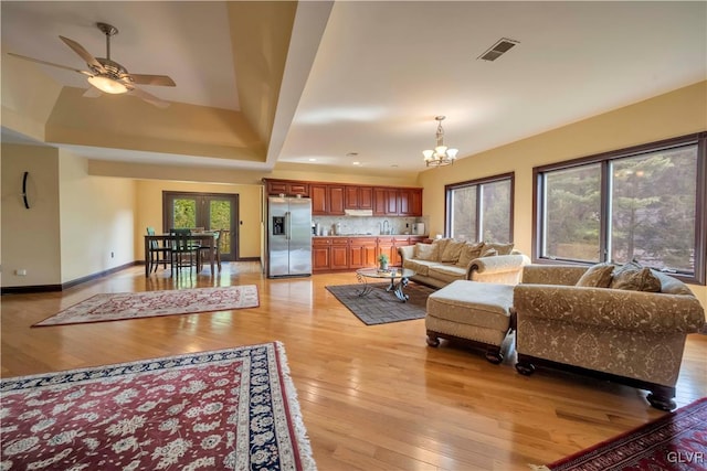 living room featuring sink, french doors, light hardwood / wood-style floors, ceiling fan with notable chandelier, and a tray ceiling