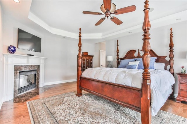 bedroom featuring a tray ceiling, a premium fireplace, light hardwood / wood-style floors, and ceiling fan