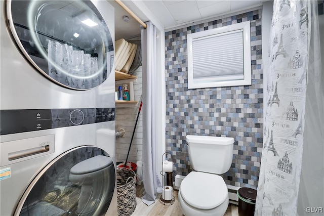 bathroom with a shower with shower curtain, a baseboard heating unit, toilet, stacked washer / dryer, and tile walls