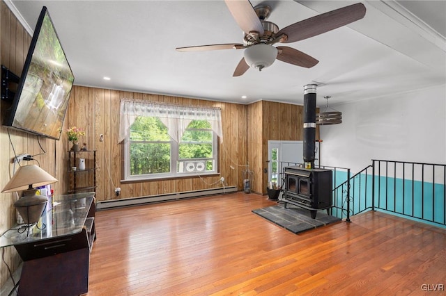 unfurnished living room featuring light hardwood / wood-style floors, baseboard heating, a wood stove, ceiling fan, and wooden walls