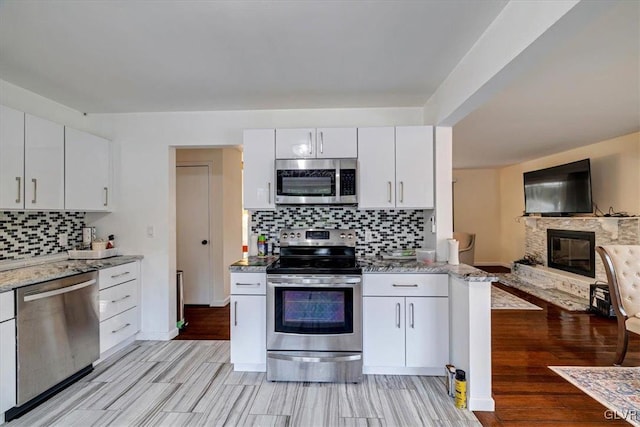 kitchen featuring light wood-type flooring, white cabinetry, stainless steel appliances, backsplash, and a stone fireplace