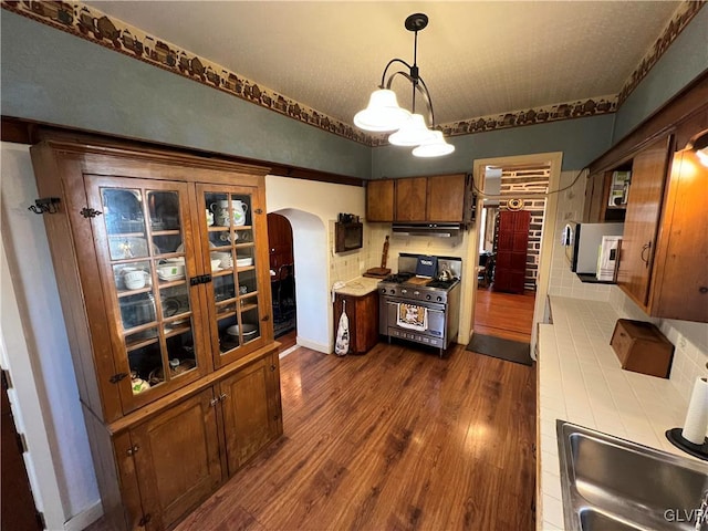 kitchen featuring brown cabinetry, dark wood finished floors, arched walkways, a sink, and stainless steel stove