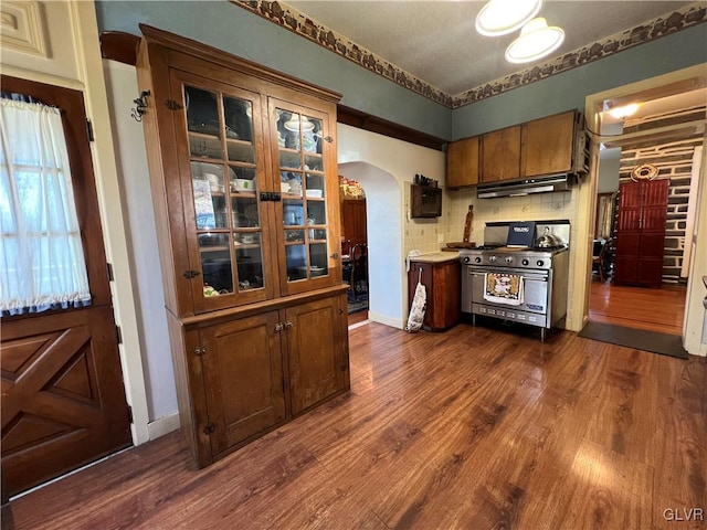 kitchen featuring dark wood-style flooring, arched walkways, decorative backsplash, under cabinet range hood, and high end stainless steel range oven