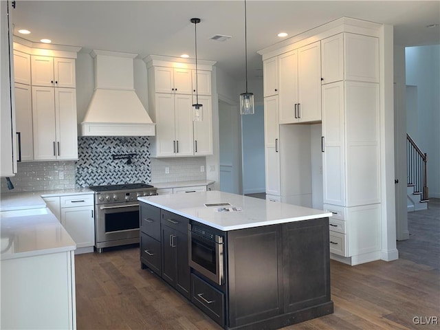 kitchen featuring white cabinetry, custom range hood, appliances with stainless steel finishes, and a kitchen island with sink