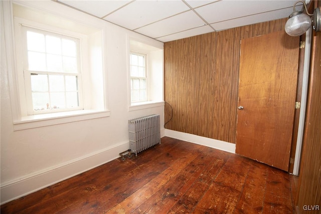 unfurnished room featuring radiator heating unit, wooden walls, a drop ceiling, and dark wood-type flooring