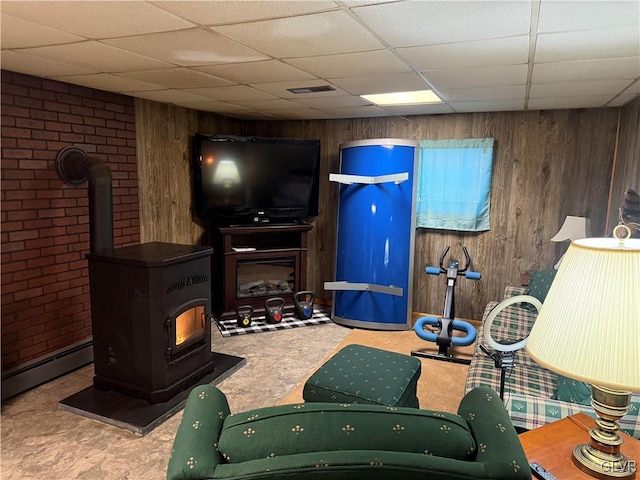 interior space with visible vents, a drop ceiling, a wood stove, wood walls, and a baseboard heating unit