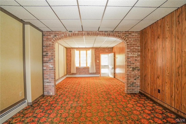 carpeted spare room with a drop ceiling, wood walls, brick wall, and a baseboard heating unit