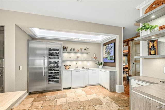 bar with stainless steel built in refrigerator, a tray ceiling, white cabinetry, and sink