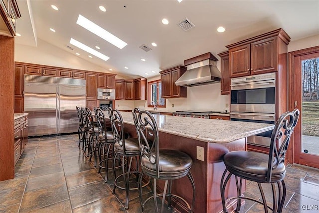 kitchen with a kitchen bar, stainless steel appliances, lofted ceiling with skylight, a spacious island, and wall chimney range hood