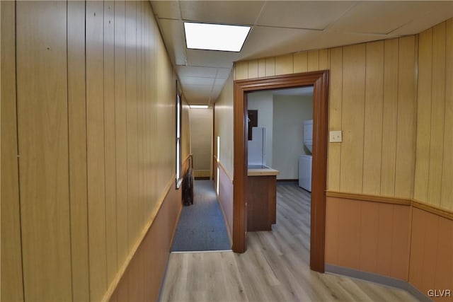 corridor with a drop ceiling, wood walls, stacked washer / dryer, and light hardwood / wood-style flooring