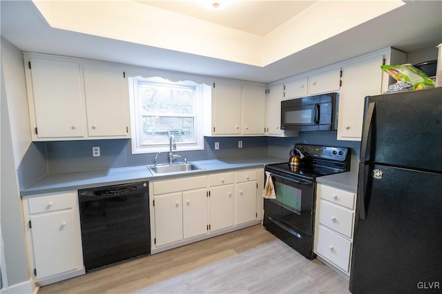 kitchen with sink, white cabinetry, a tray ceiling, black appliances, and light wood-type flooring