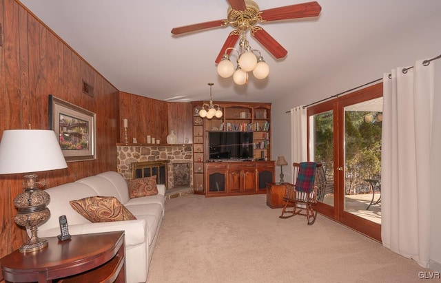 carpeted living room with a stone fireplace, wood walls, french doors, and ceiling fan with notable chandelier