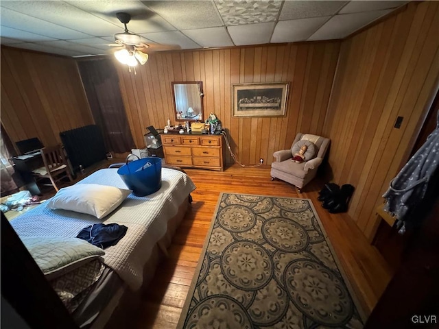 bedroom featuring ceiling fan, a drop ceiling, hardwood / wood-style floors, and wooden walls