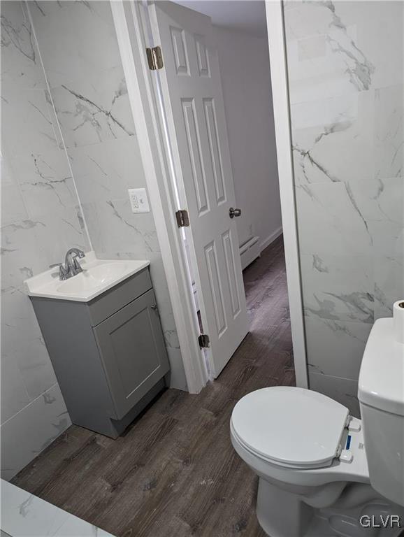 bathroom featuring a baseboard heating unit, wood-type flooring, toilet, vanity, and tile walls