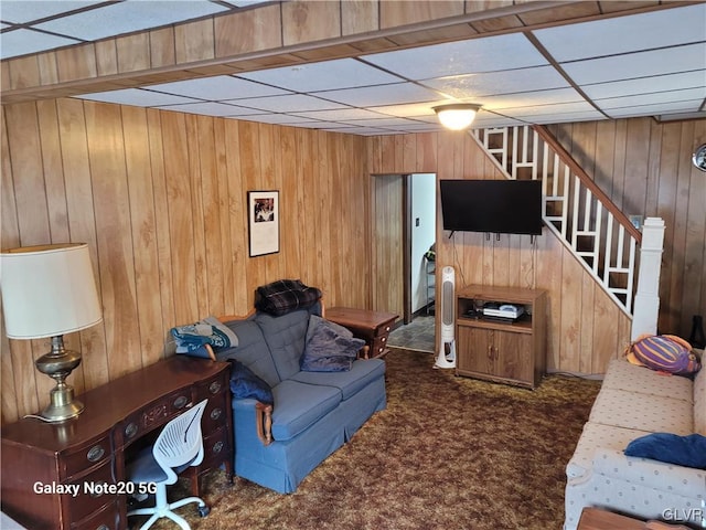 living room featuring a drop ceiling, wooden walls, and dark colored carpet