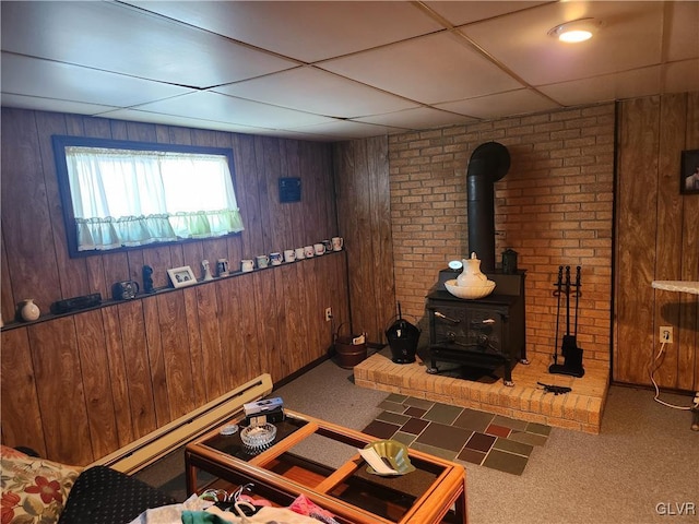 carpeted living room featuring a paneled ceiling, a wood stove, wood walls, and a baseboard radiator