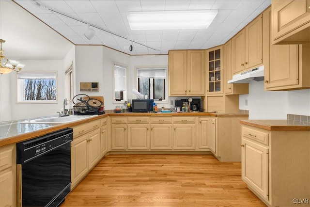 kitchen with tile countertops, under cabinet range hood, a sink, black dishwasher, and glass insert cabinets