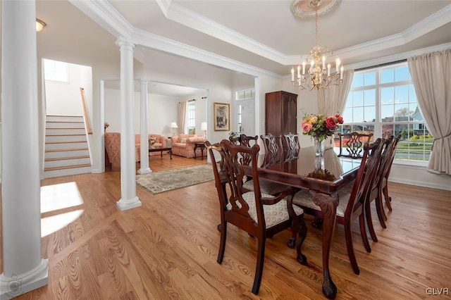 dining room with crown molding, a raised ceiling, light wood-type flooring, a notable chandelier, and decorative columns