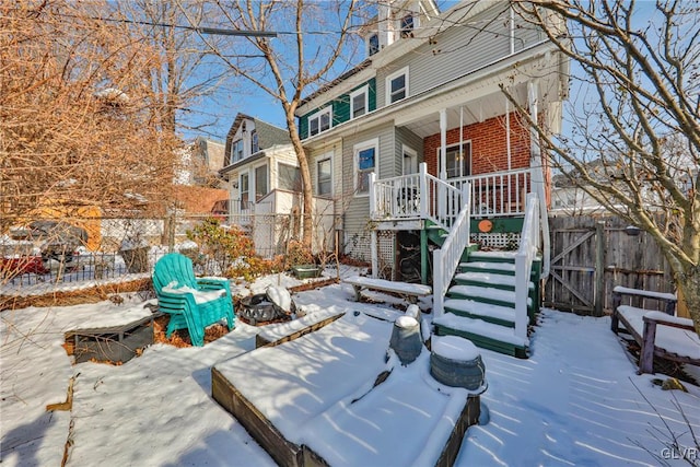 snow covered rear of property featuring a porch, brick siding, fence, and stairway