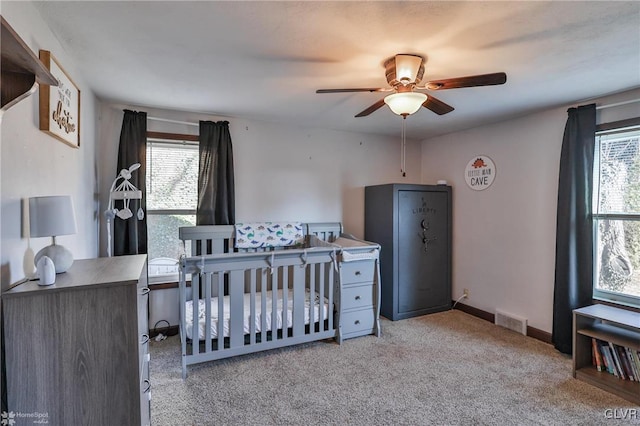 bedroom featuring multiple windows, light colored carpet, a nursery area, and ceiling fan