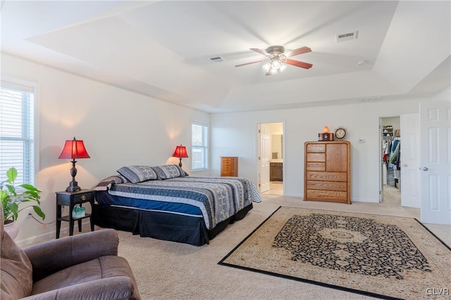 carpeted bedroom with a walk in closet, connected bathroom, ceiling fan, and a tray ceiling