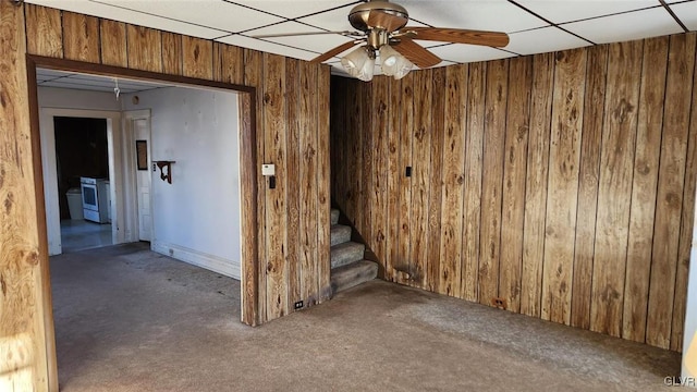 empty room with a drop ceiling, dark colored carpet, ceiling fan, and wood walls